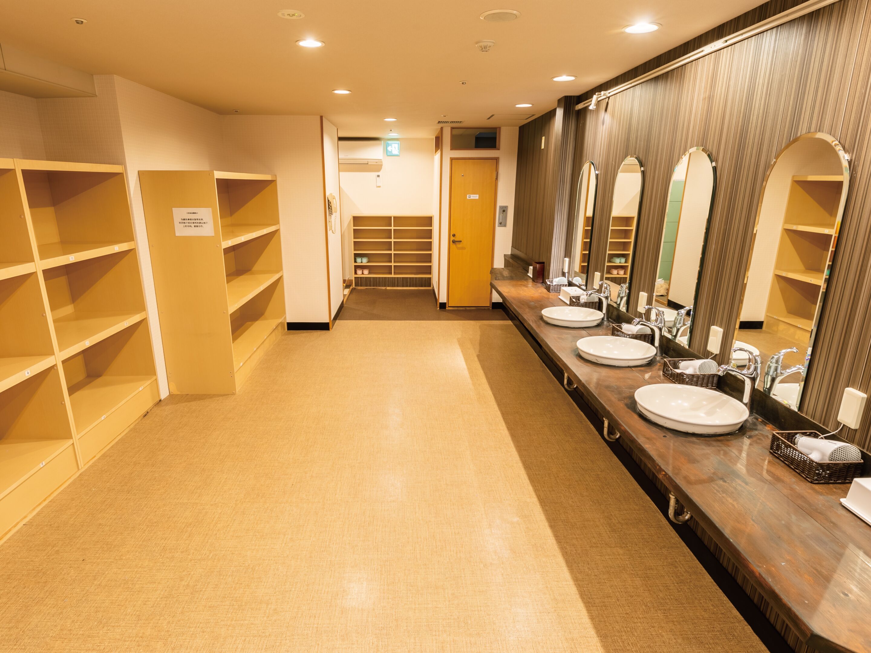 Une longue et étroite salle de bain avec plusieurs lavabos et miroirs. Des étagères se trouvent sur le côté gauche, et une porte se trouve au fond de la pièce.