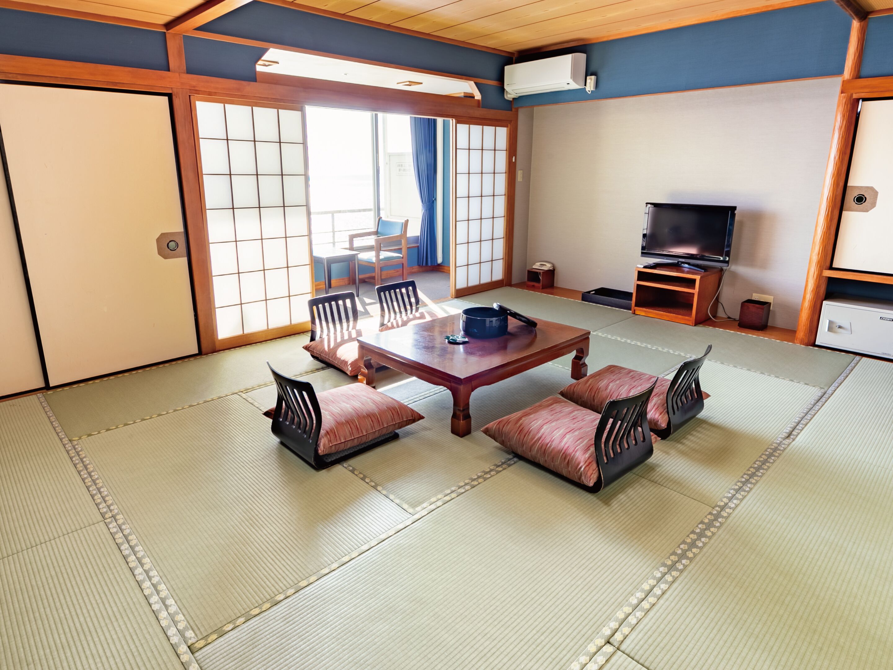 A traditional Japanese room with tatami flooring, a low wooden table with cushions, a television, and sliding doors.