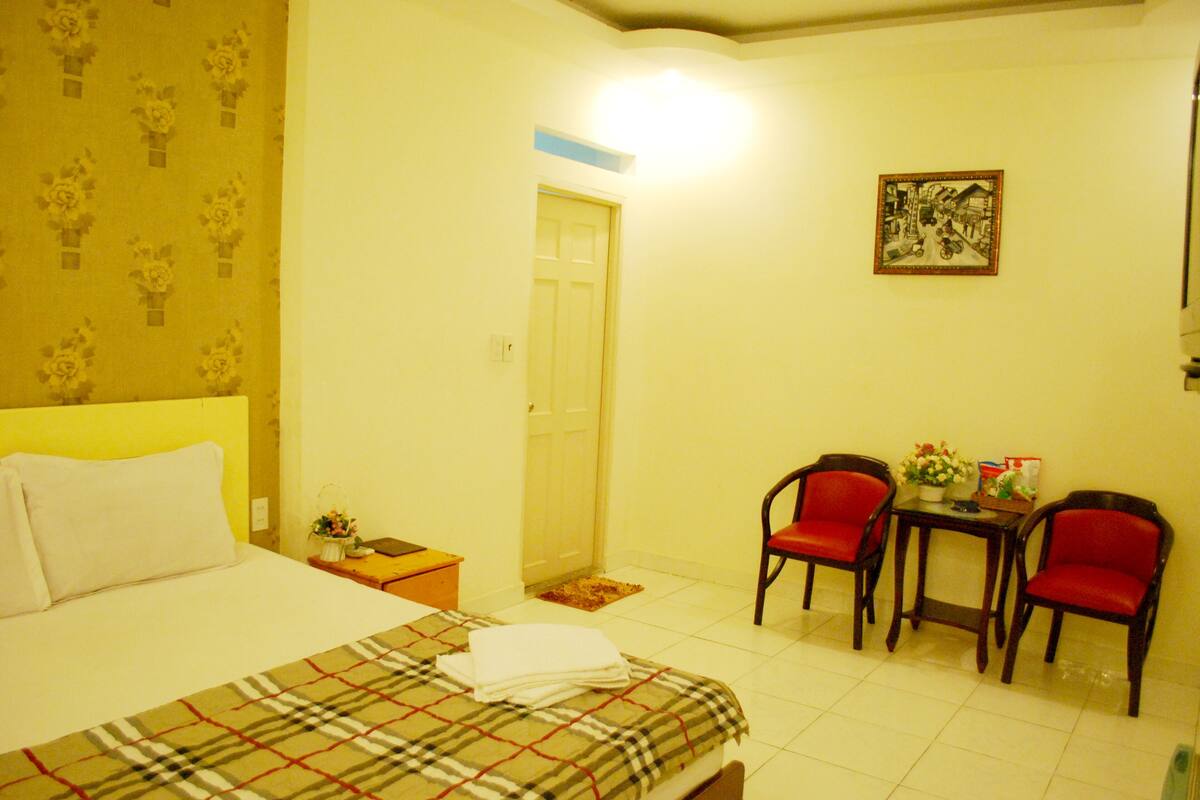 A hotel room with a bed, a nightstand, a chair, a small table, and a wall-mounted picture.