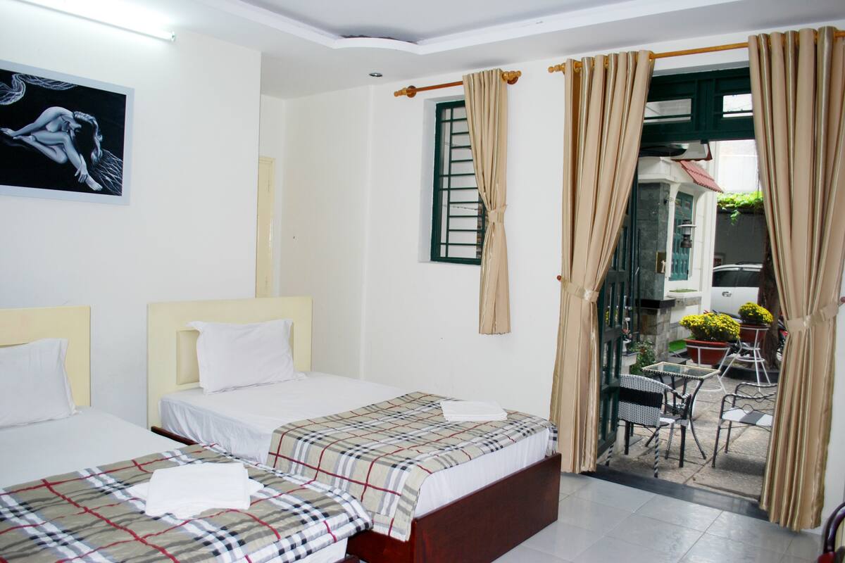 A hotel room with two beds, a window with curtains, a small balcony with outdoor furniture, and a painting on the wall.