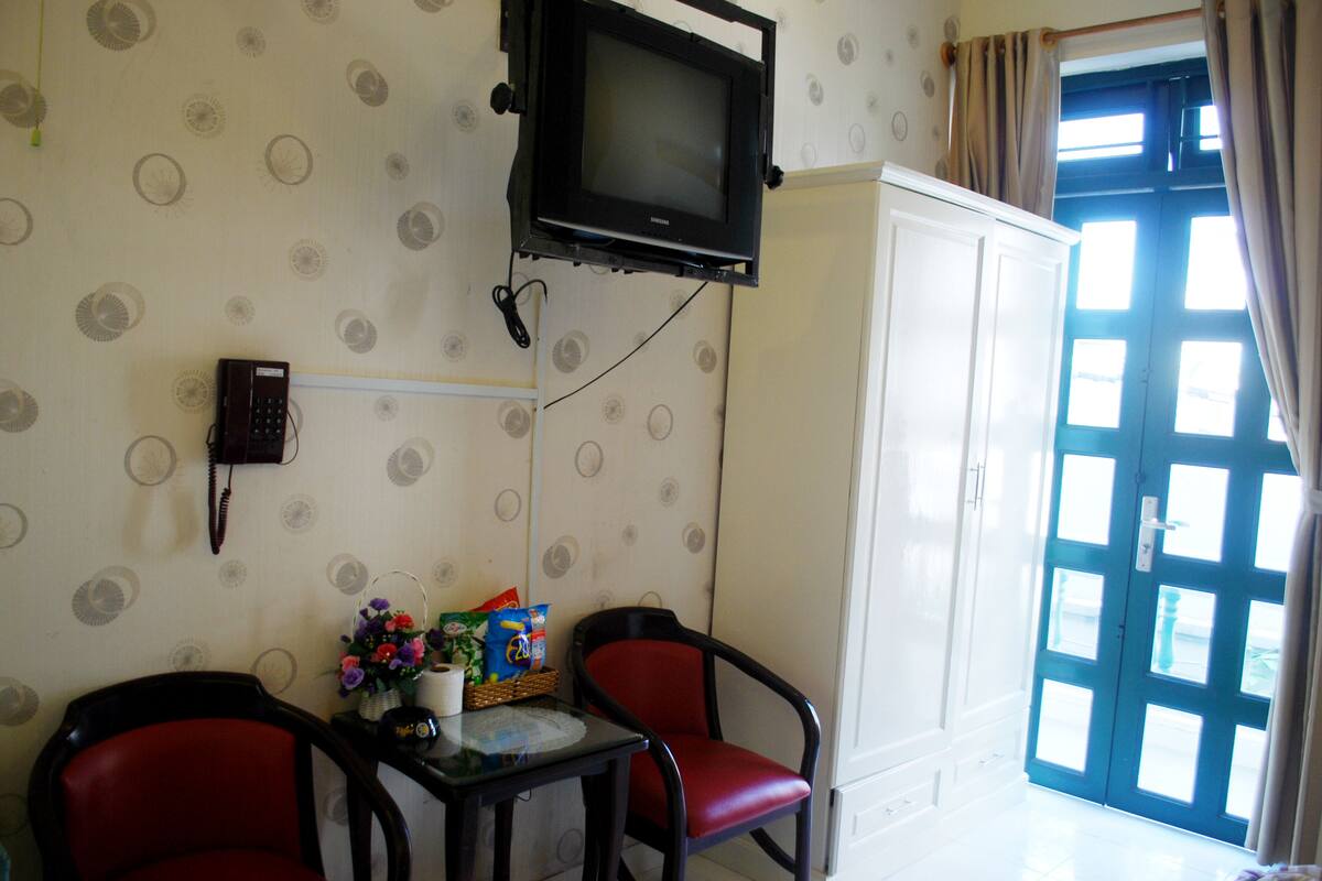 A room with a television mounted on the wall, a telephone, two red chairs, a small table with a basket of flowers, and a white cabinet.