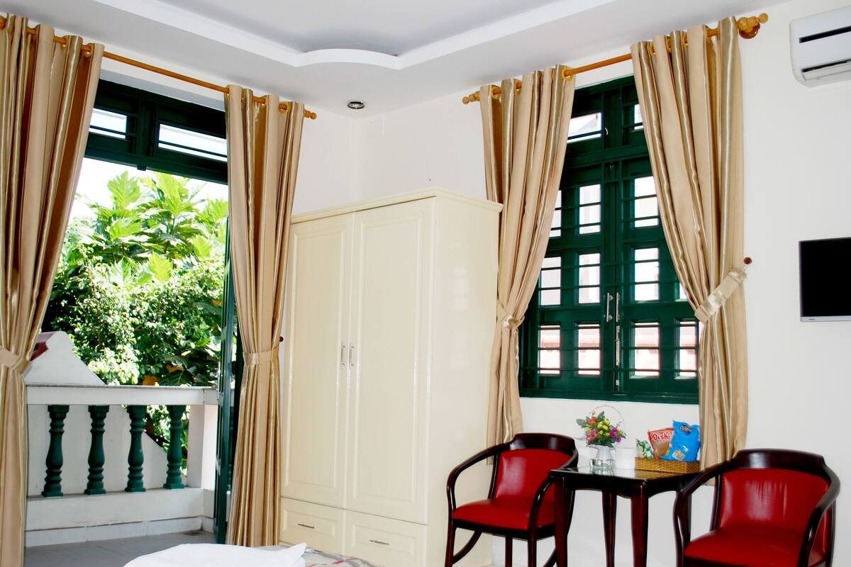 A room with a white wardrobe, two red chairs, a table with a basket and snacks, and a window with green shutters.