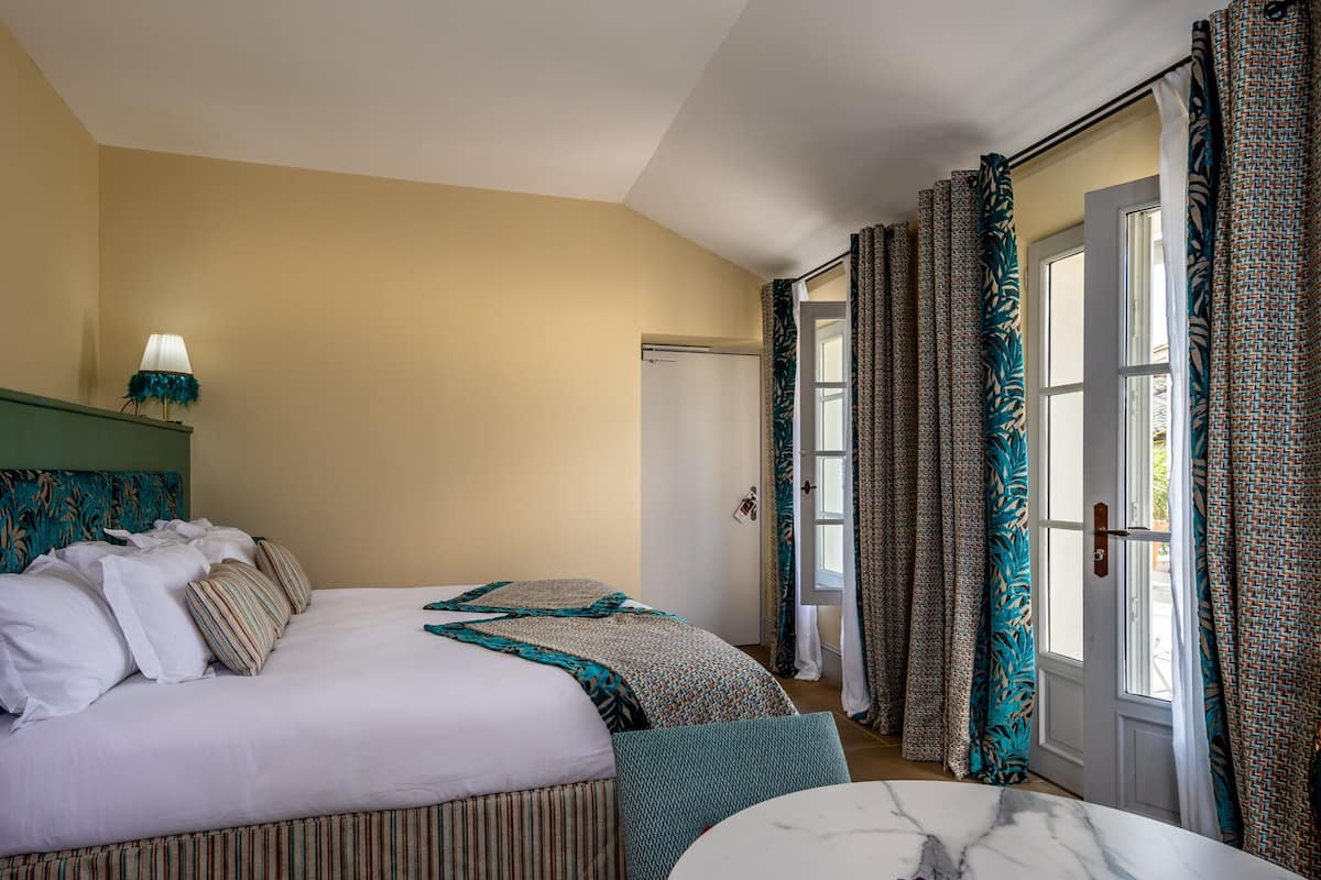 A hotel room with a bed, a nightstand, a lamp, a window with curtains, and a door.
