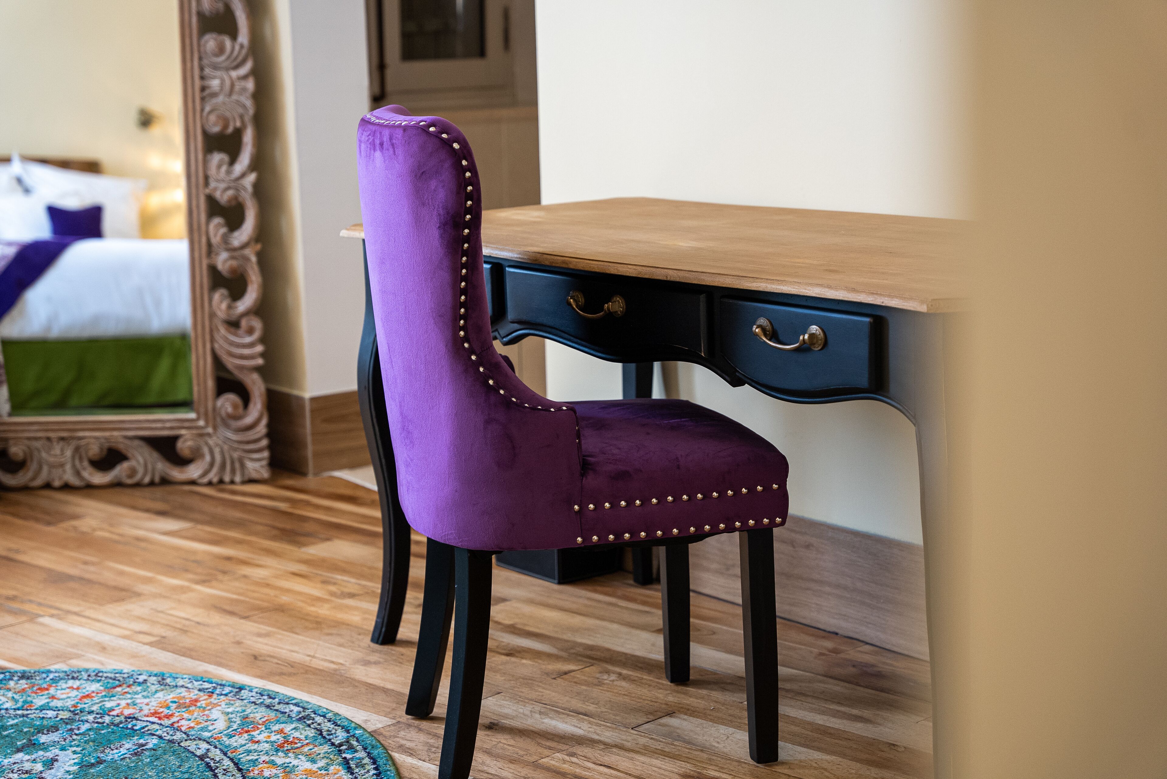 A purple upholstered chair with gold studs, a wooden desk with two drawers, and a patterned rug on a wooden floor.