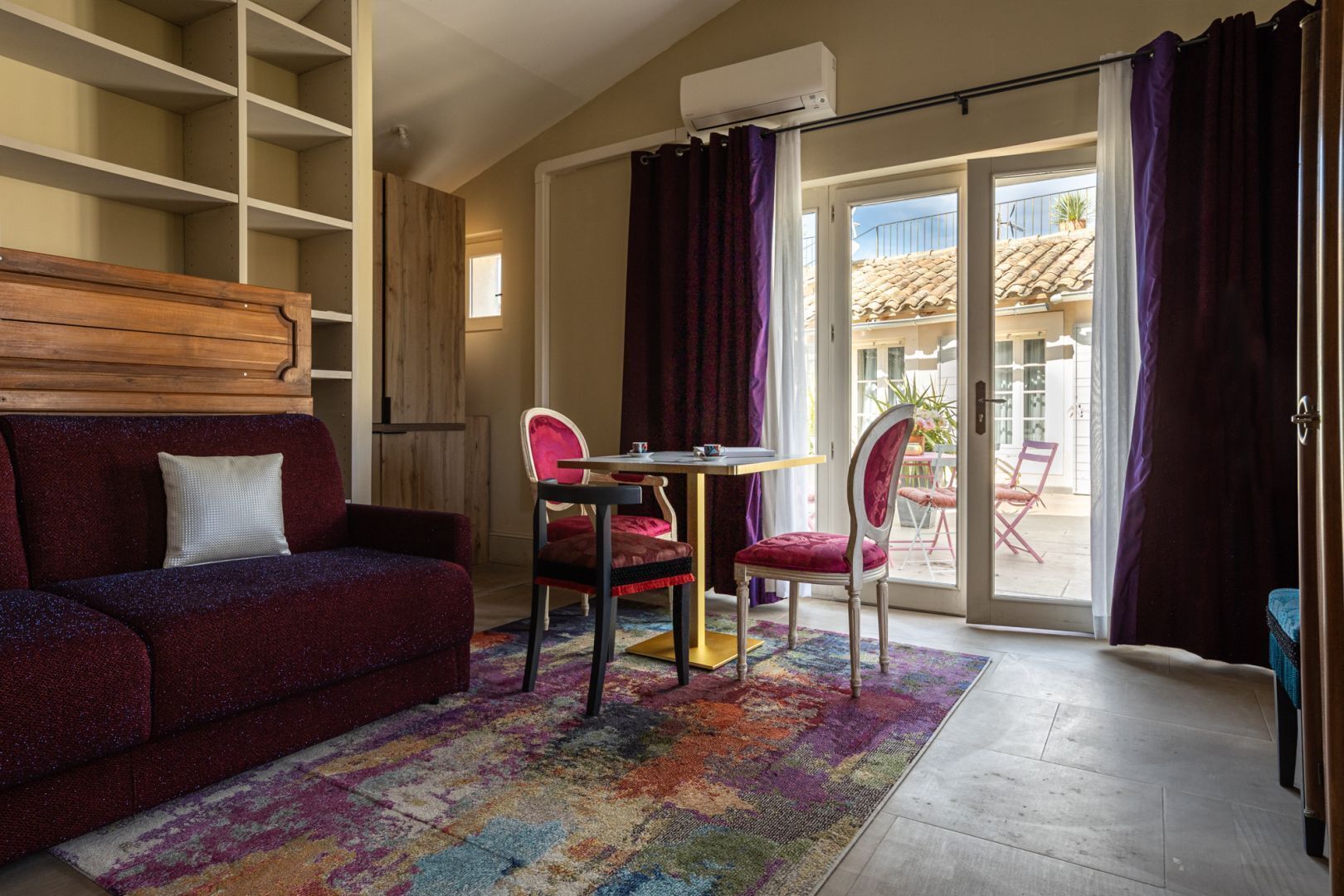 Un salon chaleureux avec un canapé, deux fauteuils, une petite table et un tapis coloré. Une porte-fenêtre coulissante donne accès à un espace extérieur d’où l’on peut admirer un bâtiment dont le toit est recouvert de tuiles.