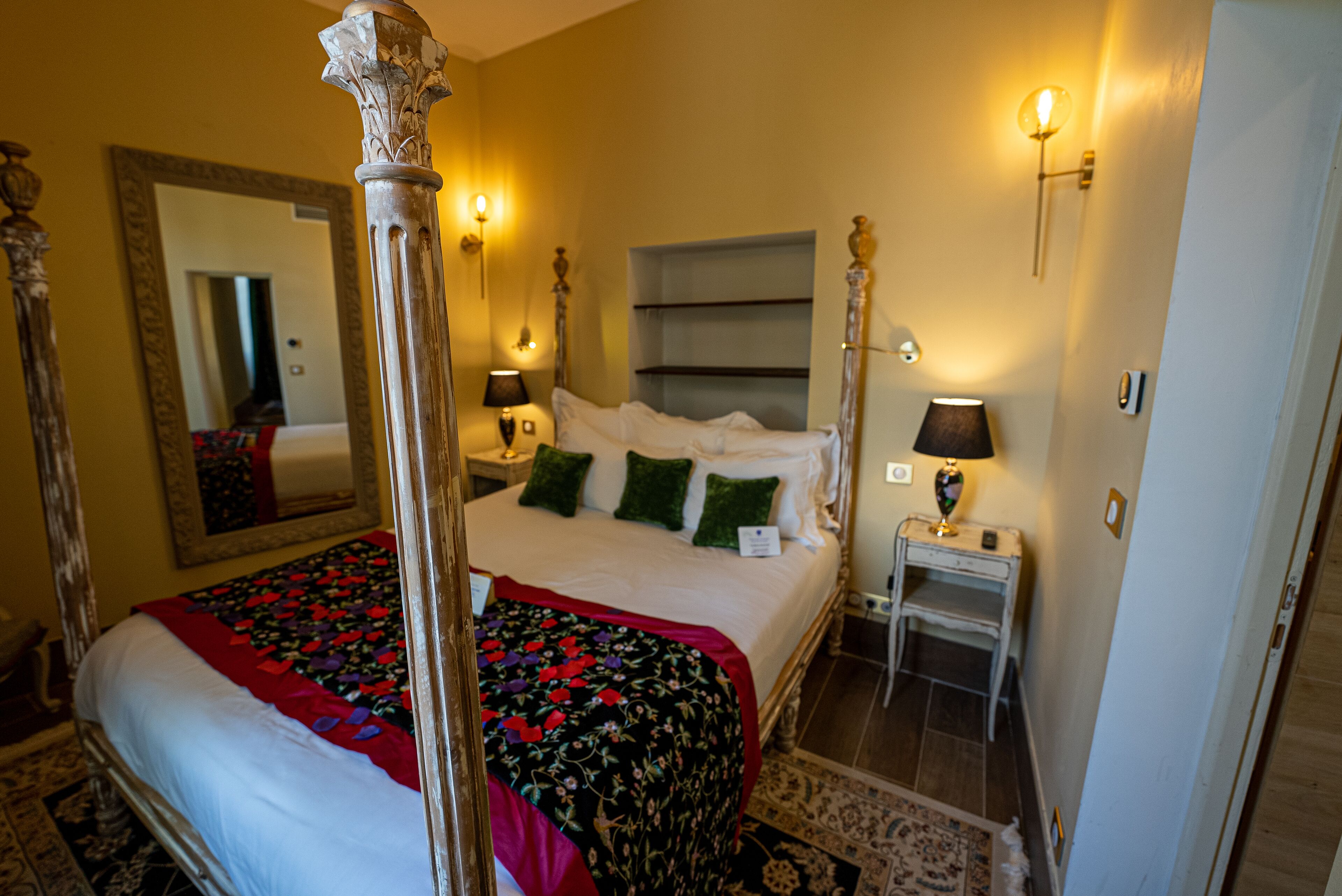 A four-poster bed with a floral bedspread, two bedside tables with lamps, a mirror, and a nightstand.