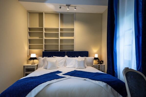 A hotel room with a large bed, two bedside tables, a wardrobe with multiple shelves, and a window with blue curtains. - Le Clos Violette D'Aglaé (L'Isle-sur-la-Sorgue)