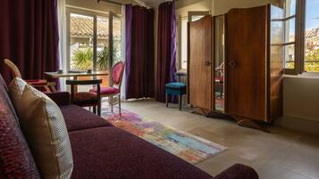 A cozy living room with a sofa, a table, and a chair. There is a wooden wardrobe and a window with curtains.