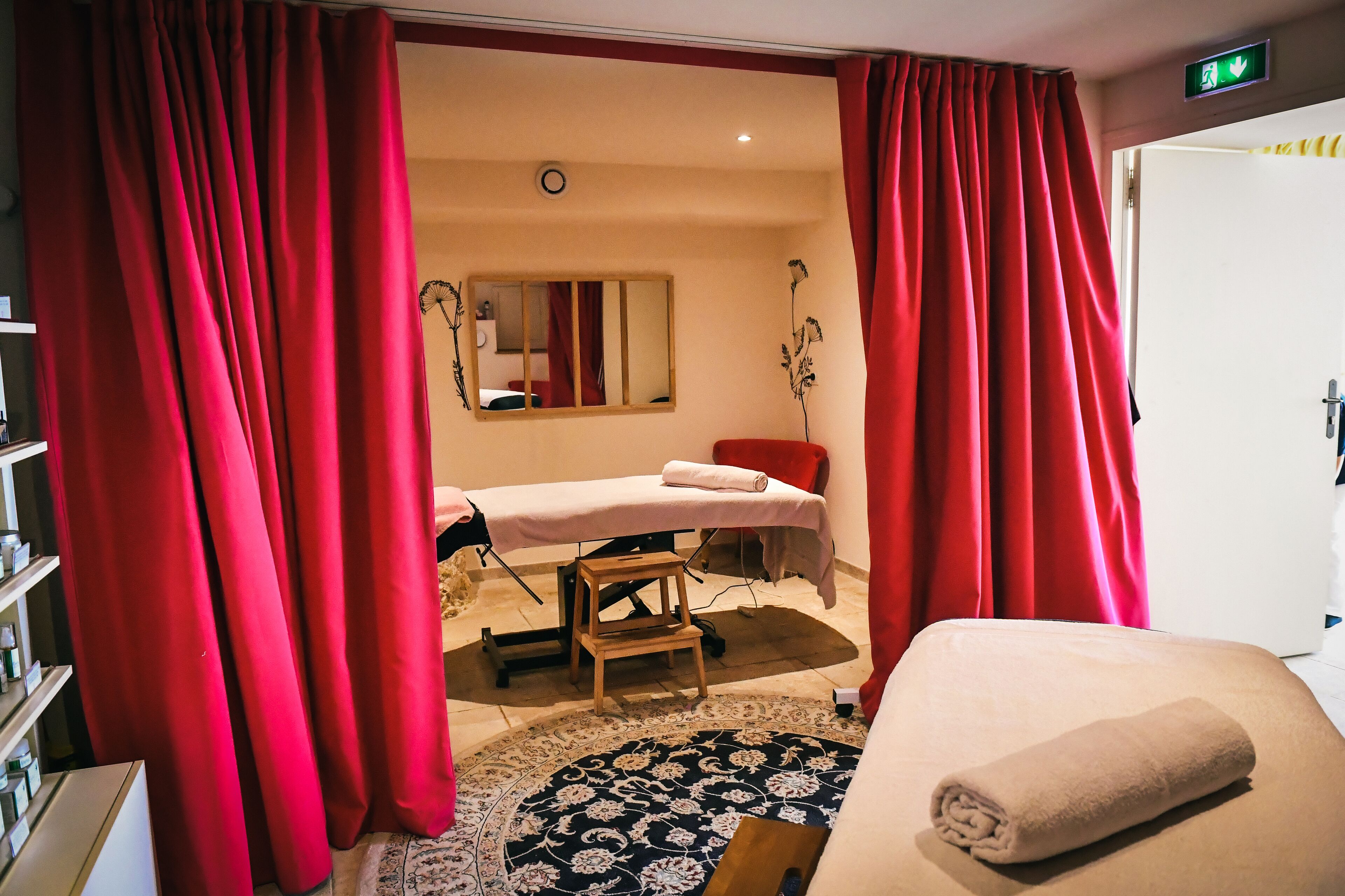 A room with a massage table, a small wooden stool, a patterned rug, and a rolled towel on a bed.