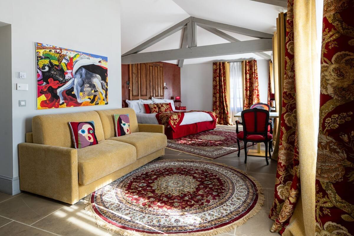 A spacious room with a large bed, a sofa, and a dining area. There is a painting on the wall and a window with curtains.