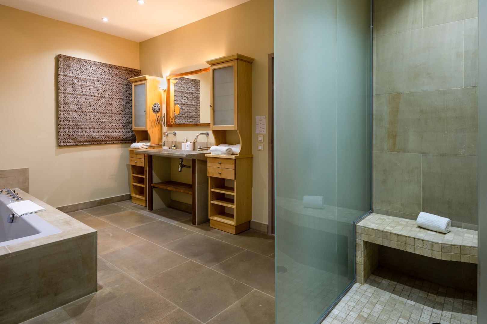 Une salle de bain dotée d’une grande baignoire, de deux vasques avec des meubles en bois, et d’une douche séparée par des parois en verre.