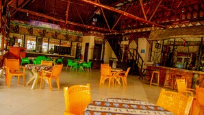 Restaurante com teto alto e vigas aparentes, com cadeiras de plástico verde e amarelo e uma mesa com toalha estampada.