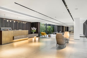 A modern reception area with a wooden reception desk, seating with cushions, and a view of greenery outside. - Luxury with Terrace & Sea View by FeelHome (Tel Aviv)