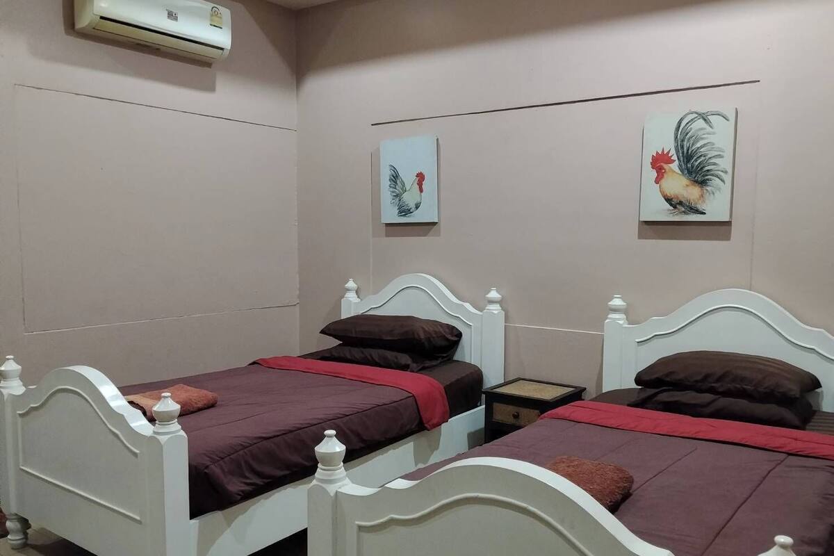 Une chambre à coucher avec deux lits blancs, chacun recouvert d’une couverture rouge et agrémenté de coussins marron. Deux tableaux sont accrochés au mur, l’un représentant un coq et l’autre un oiseau. Un appareil de climatisation est fixé au mur, au-dessus des lits.