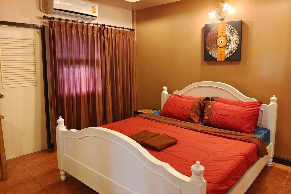 Une chambre à coucher bien rangée, avec un lit blanc, une literie rouge et marron, une œuvre d’art accrochée au mur et une porte fermée.