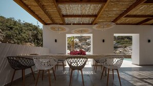 Un área de comedor al aire libre con una mesa y sillas de madera, rodeada por un techo y paredes de bambú, con vista a un paisaje rocoso.