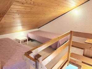 A small, single-room attic bedroom with a sloped ceiling, a bunk bed, a bedside table, and a lamp.