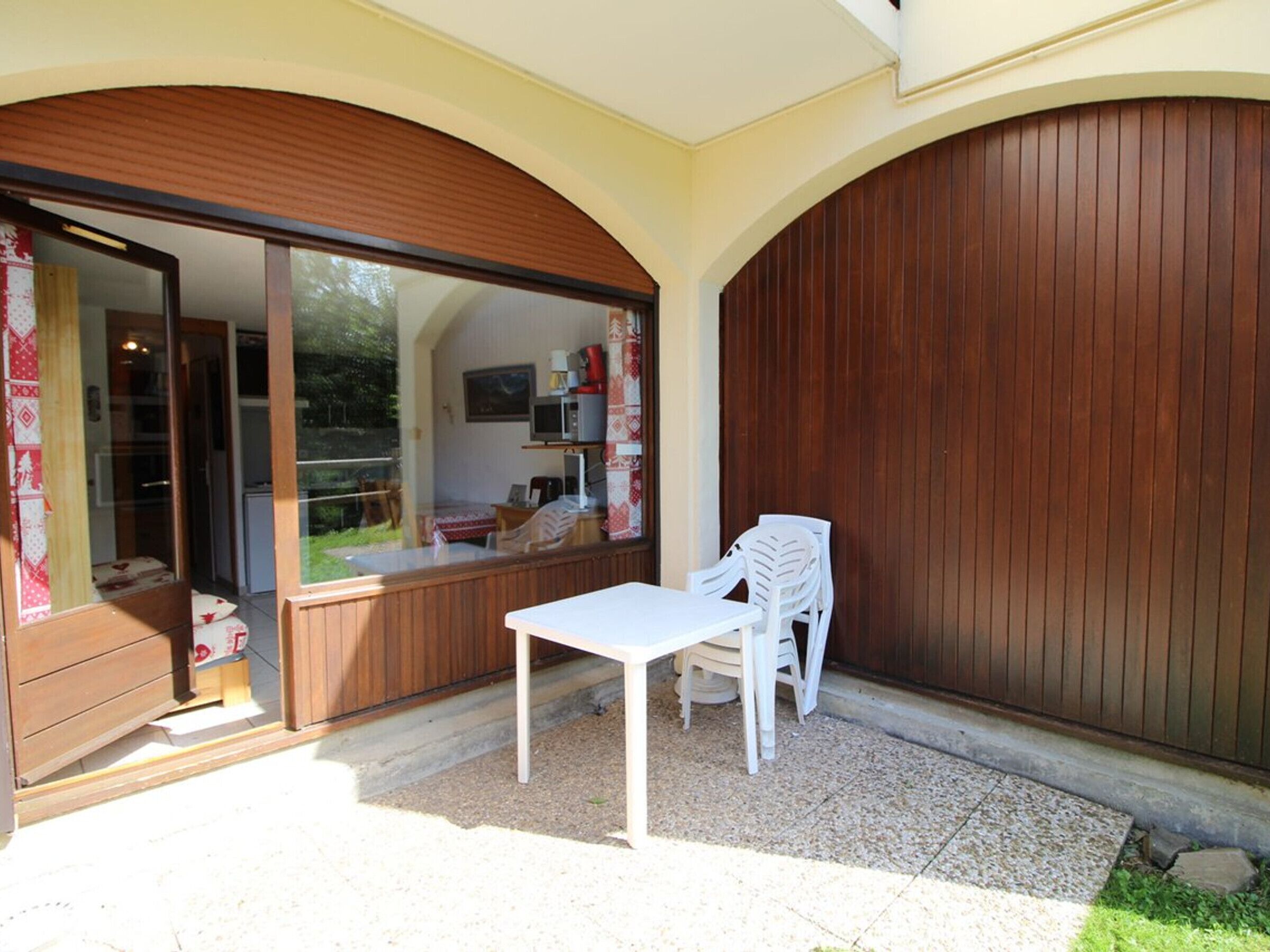 Un espace patio extérieur avec une table et des chaises blanches, une clôture en bois et une vue sur l’intérieur de la maison à travers une grande porte vitrée.