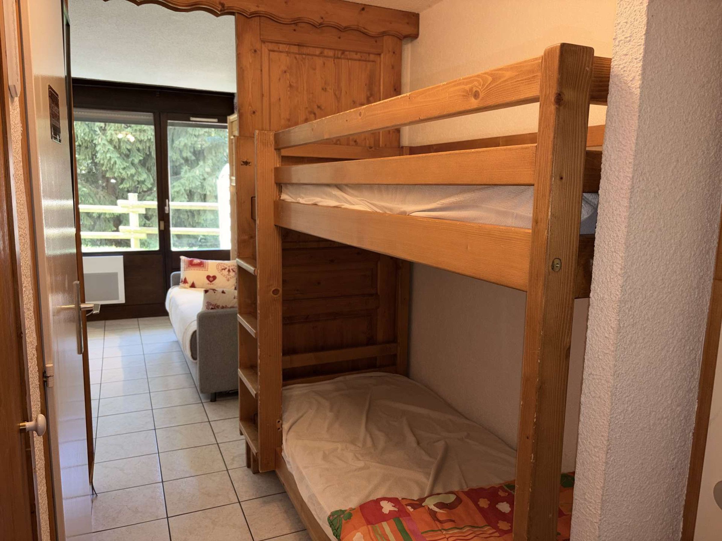 A bunk bed with a wooden frame, a single bed with a patterned bedspread, and a window with a view of greenery.