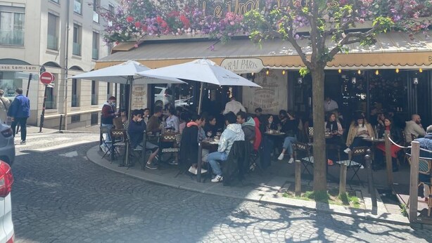 Um café ao ar livre com mesas e guarda-sóis, rodeado por edifícios e uma árvore com flores cor-de-rosa.
