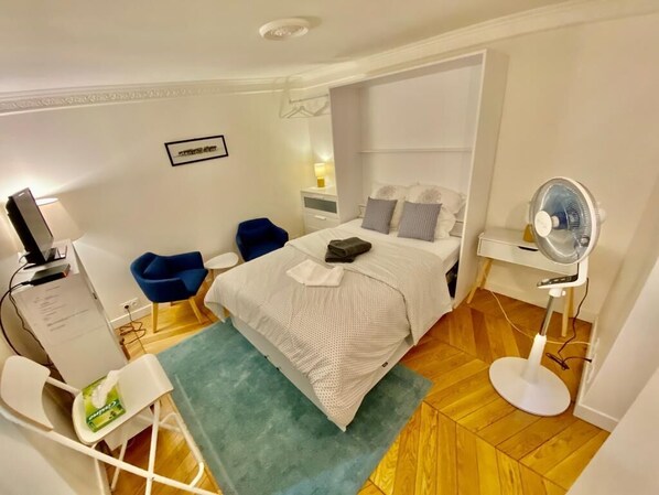 A bedroom with a bed, a desk, a chair, a mirror, and a fan.