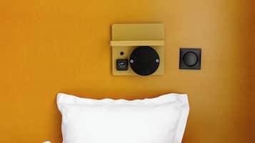 A bed with white bedding against a yellow wall, a small black speaker, and a beige panel with a circular object and a USB port.
