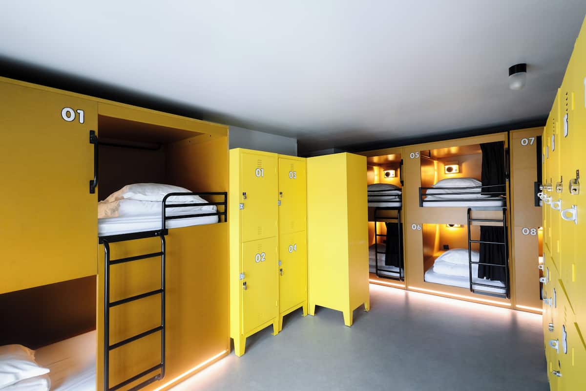 Ein Schlafsaalzimmer mit Stockbetten, gelben Schließfächern und einem kleinen Fenster.