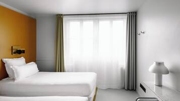 A hotel room with two beds, a window with curtains, a bedside table with a lamp, and a wall-mounted light fixture.