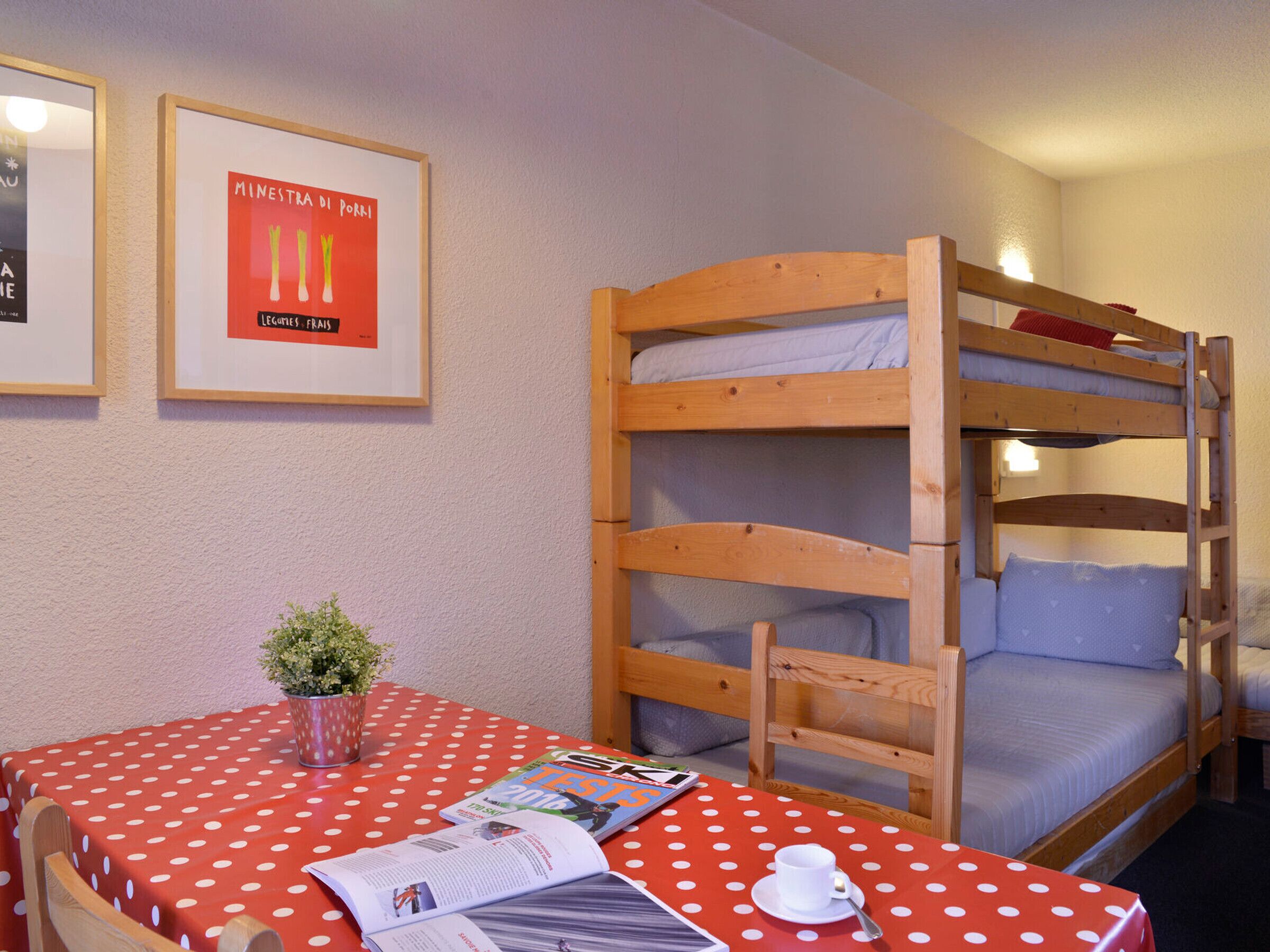 Une chambre avec un lit superposé, une nappe à pois, un menu encadré et une petite plante en pot.
