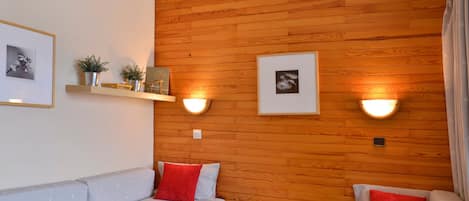A cozy room with a wooden wall, two beds with red pillows, a small wooden nightstand, and wall-mounted lights.