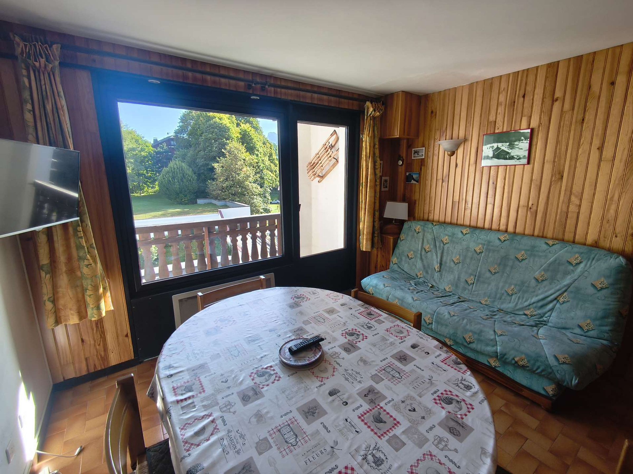 A room with a round table, a sofa, a TV, and a balcony with a view of greenery.