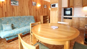 Ein gemütliches Zimmer mit einer Holzwand, einem blau gemusterten Sofa, einem Holzesstisch mit Stühlen und einer Küchenzeile mit Holzausbau und Geräten.