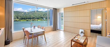 Habitación de hotel moderna con un ventanal grande que ofrece vistas panorámicas de un lago y montañas, un comedor con mesa y sillas, y una zona de estar acogedora con una mesita de centro.