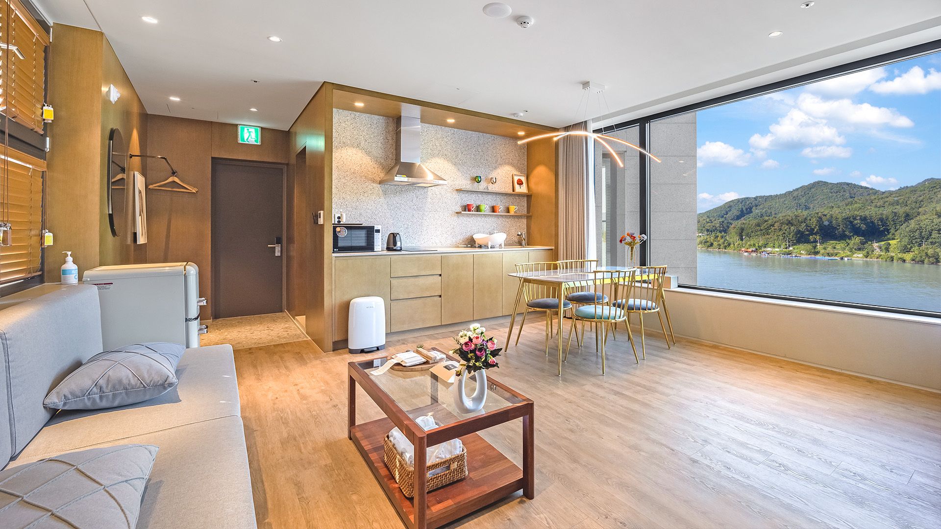 A modern hotel room with a kitchenette, a dining area with a table and chairs, a sofa, and a large window with a view of a lake and mountains.