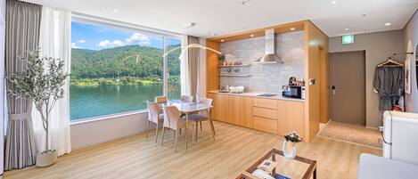 A modern hotel room with a dining area, kitchenette, and a large window offering a scenic view of a lake and hills.