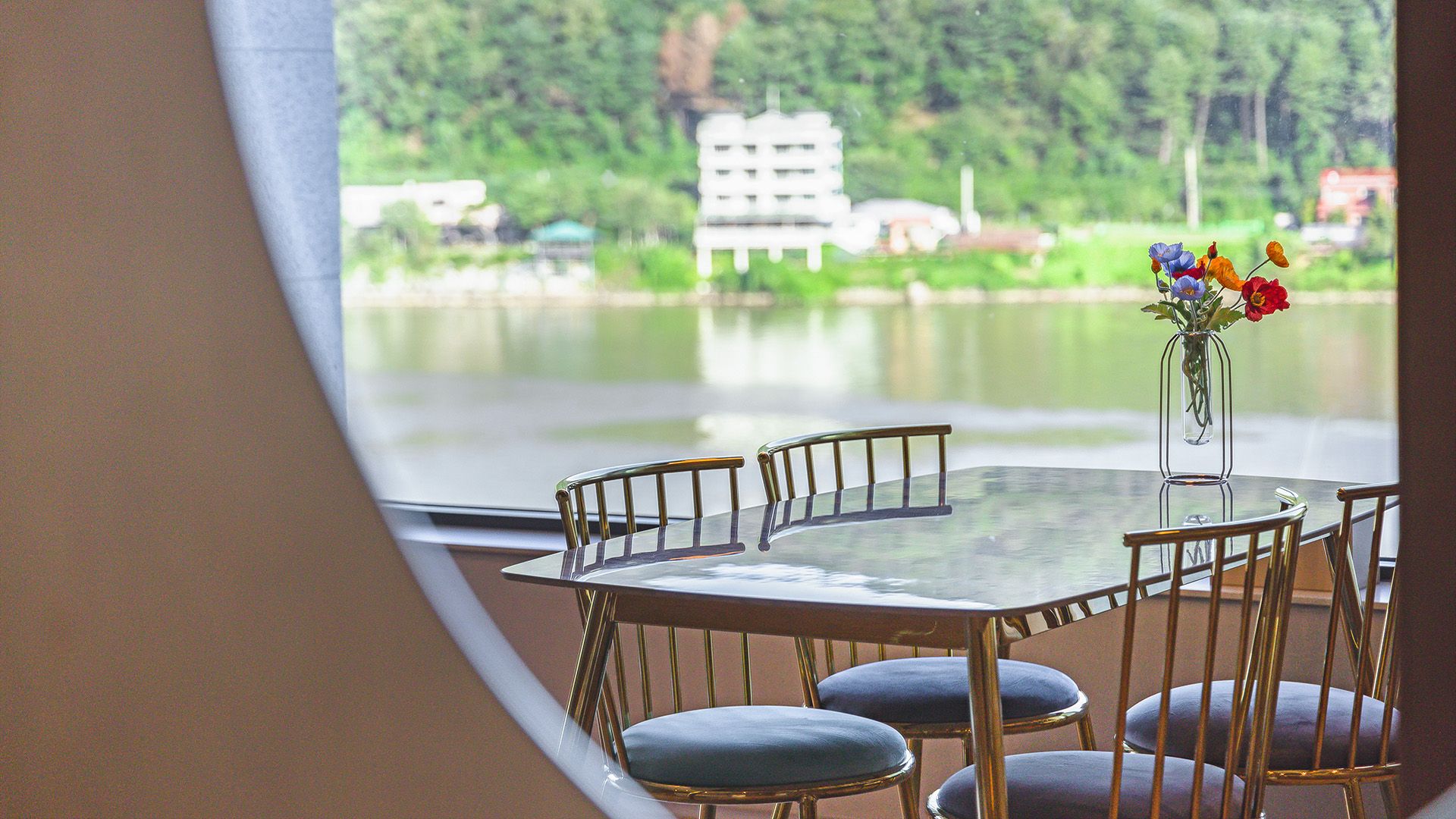 A round table with chairs, a vase with flowers, and a view of a lake with buildings in the background.