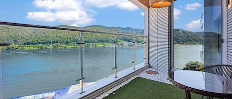 Ein Balkon mit Glasgeländer, der auf einen See blickt, mit Tisch und Stühlen und Blick auf die umliegende Landschaft.