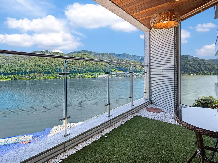 Ein Balkon mit Glasgeländer, der auf einen See blickt, mit Tisch und Stühlen und Blick auf die umliegende Landschaft.