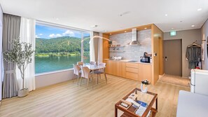 Une chambre d’hôtel moderne dotée d’un coin repas, d’une kitchenette et d’une grande fenêtre offrant une vue sur un lac et des collines.