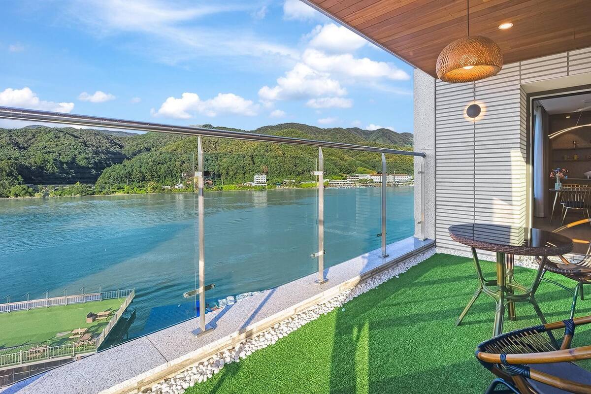 Ein Balkon mit Glasgeländer, der auf einen See blickt, mit einem Tisch und Stühlen sowie Blick auf eine Berglandschaft.