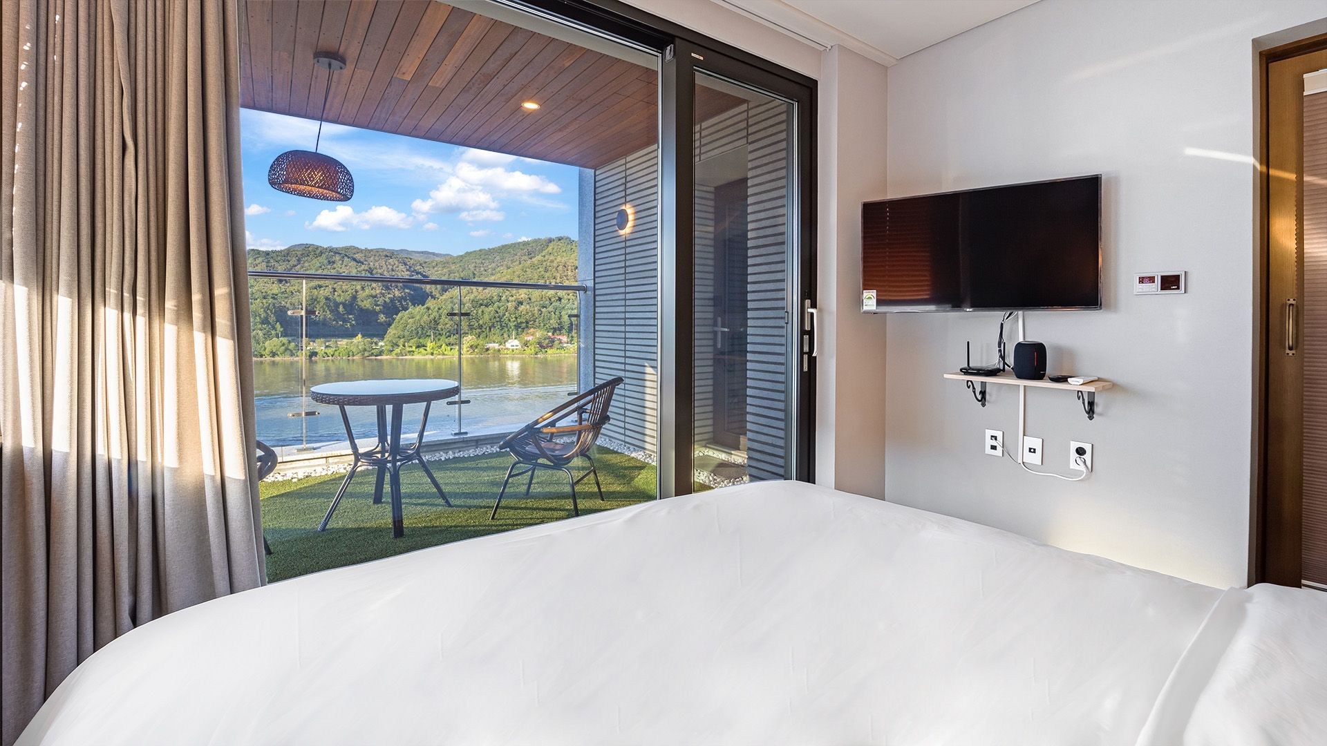 Ein Hotelzimmer mit einem Bett, einem an der Wand montierten Fernseher, einem kleinen Tisch mit Stühlen und einem Balkon mit Blick auf einen See und Hügel.