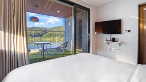 Une chambre d’hôtel avec un lit, un téléviseur fixé au mur, une petite table avec des chaises et un balcon offrant une vue sur un lac et des collines.