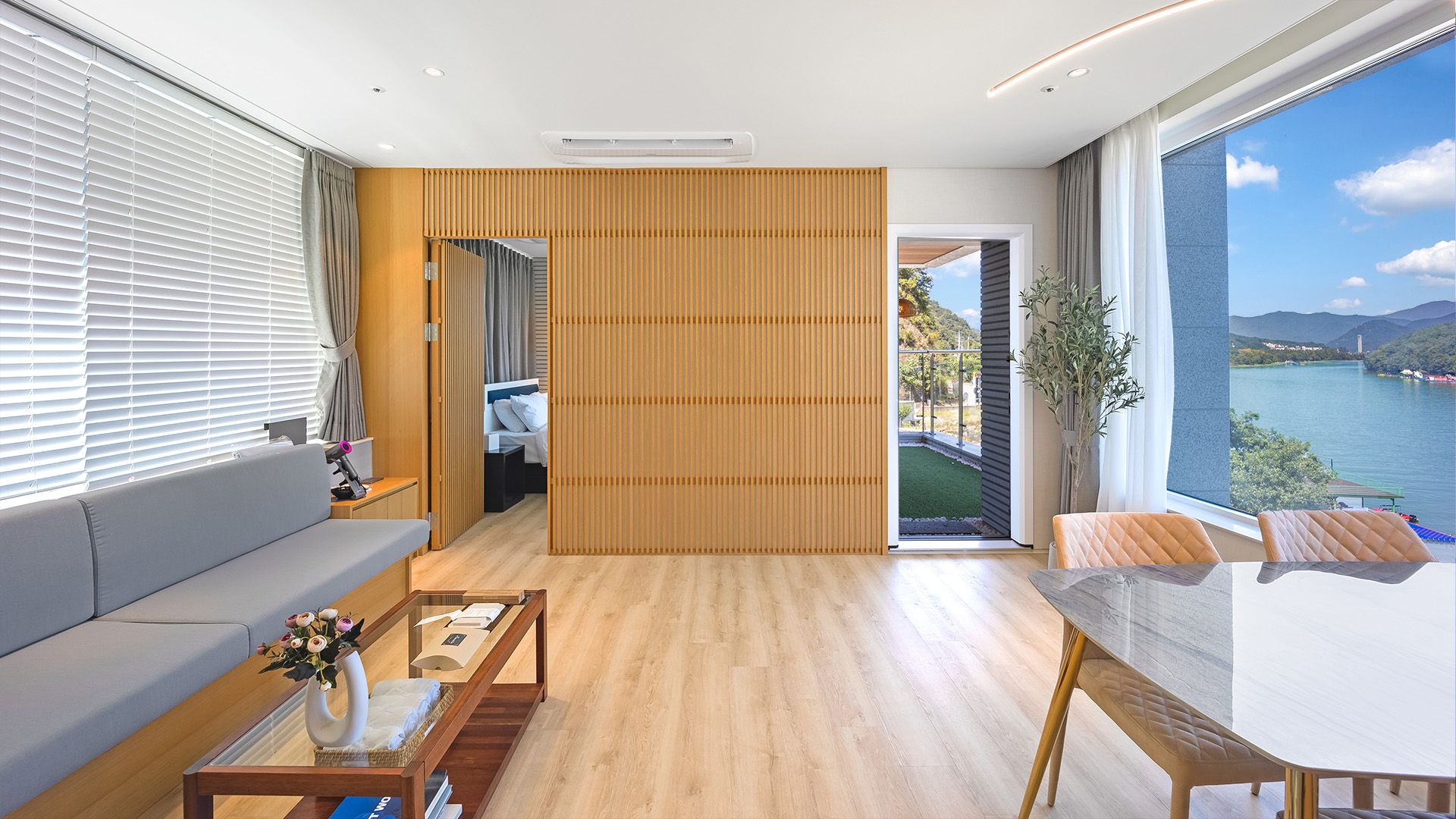 Une chambre d’hôtel moderne avec un plancher en bois, une table basse en verre, un canapé, une table à manger et une vue sur un lac.