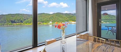 A room with a large window overlooking a lake, a vase with flowers on the table, and a view of a boat on the water.