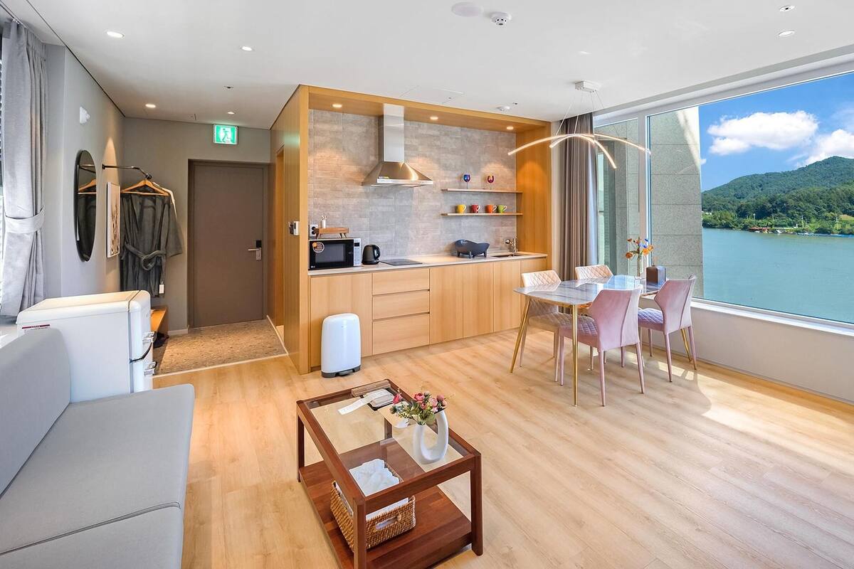 Ein modernes Hotelzimmer mit Holzboden, einer Essecke mit Tisch und Stühlen, einer Küchenzeile, einem großen Fenster mit Blick auf einen See und Berge sowie einer kleinen Sitzecke mit Tisch und Kissen.