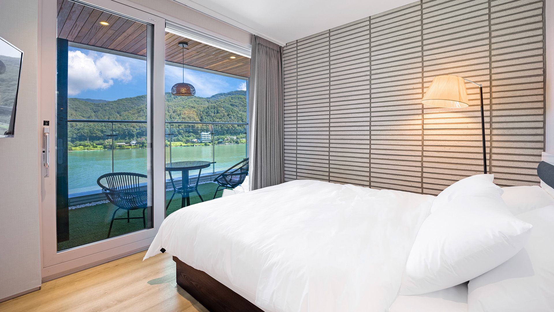 A bedroom with a large bed, a view of a lake and mountains through a sliding glass door, and a wall-mounted lamp.
