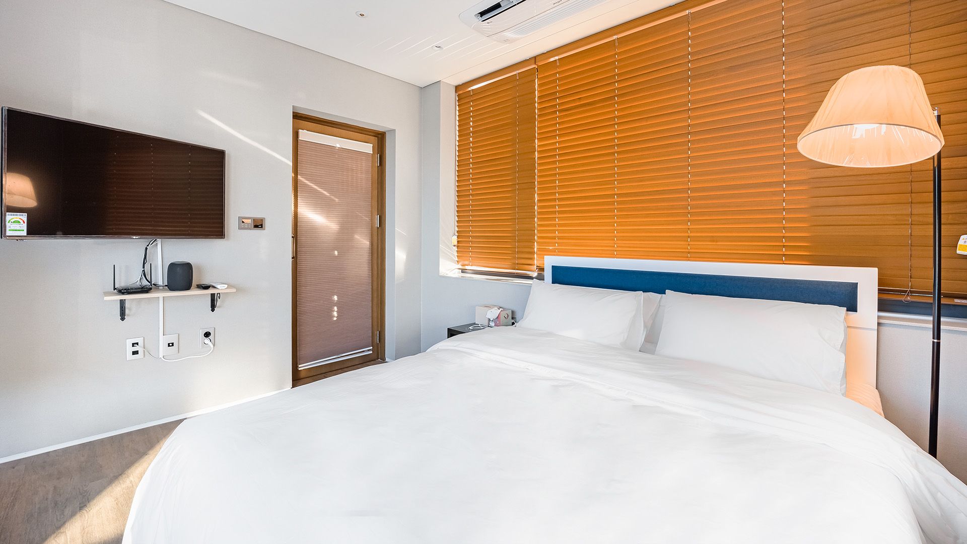 A hotel room with a bed, a TV mounted on the wall, a bedside table with a lamp, and a window with blinds.