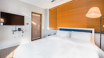 Habitación de hotel con cama, televisor montado en la pared, mesita de noche con lámpara y ventana con persianas.