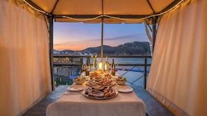 Mesa de comedor dispuesta bajo una carpa, con vista a una masa de agua y colinas al fondo.