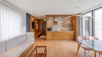 A modern hotel room with a kitchenette, a seating area with a sofa and a glass table, and a large window providing a view.